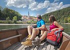 Zillenfahrt am Kloster Weltenburg Paar sitzt in einem Boot auf einem Fluss und blickt auf ein Gebäude mit Turm und Wald im Hintergrund.