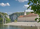 Kloster Weltenburg am Donaudurchbruch Flussufer mit Kiesstrand, altes Gebäude mit Turm und bewaldete Felsen unter blauem Himmel mit Wolken