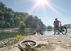 Radler an der Donaufähre bei Weltenburg Personen mit Fahrrädern stehen an einem Flussufer mit bewaldeten Felsen im Hintergrund bei Sonnenschein.
