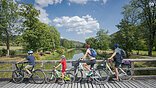 Vier Personen mit Fahrrädern auf einer Holzbrücke mit Blick auf einen Fluss und grüne Landschaft.