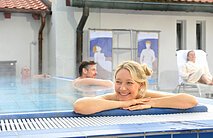 Frau lehnt entspannt am Beckenrand eines Hallenbads, im Hintergrund zwei Männer im Wasser und eine Frau auf Liegestuhl.